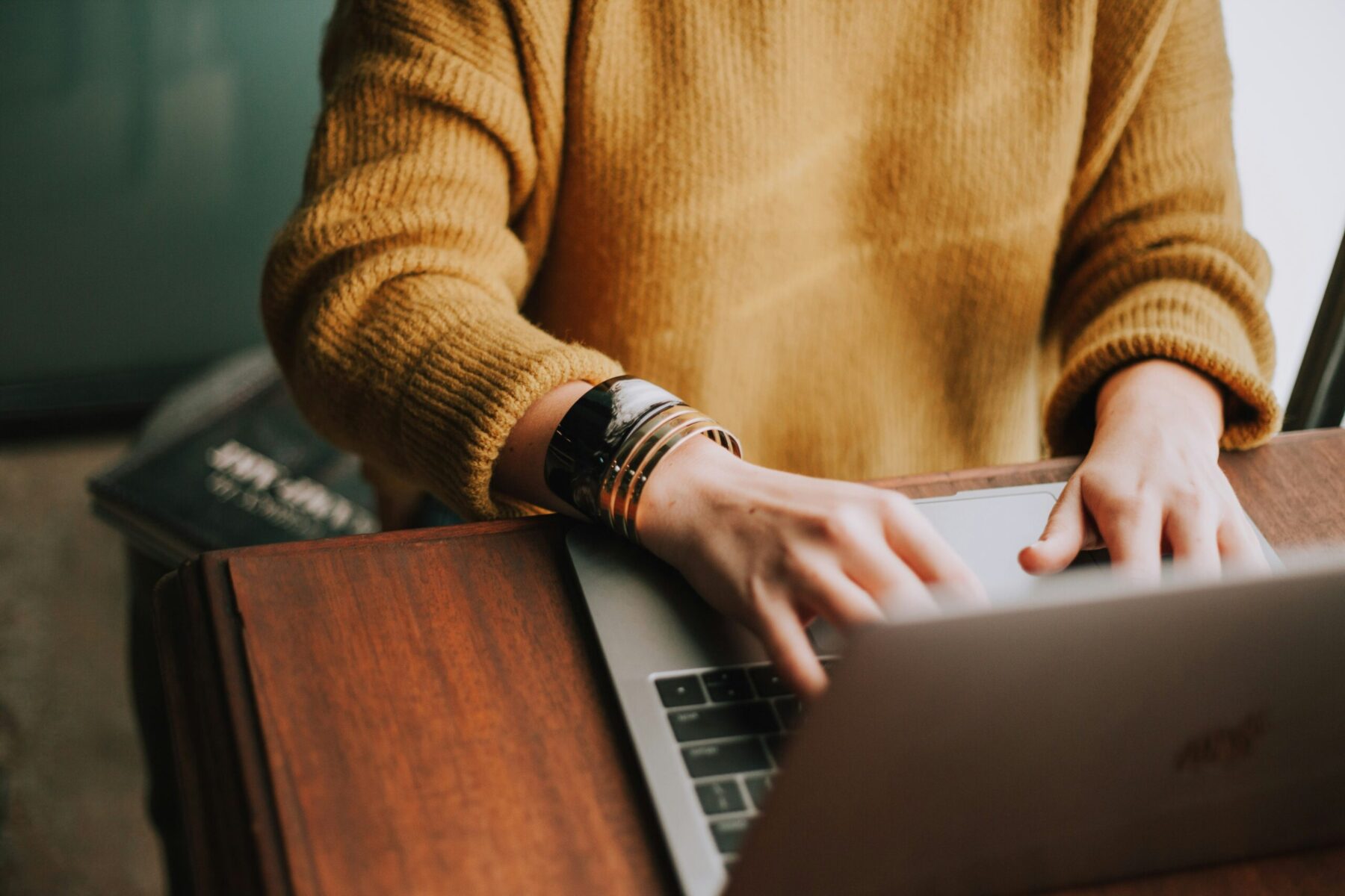 christin-hume-Hcfwew744z4-unsplash The image shows a person sitting at their desk with their hands on their laptop. They are wearing a yellow jumper.